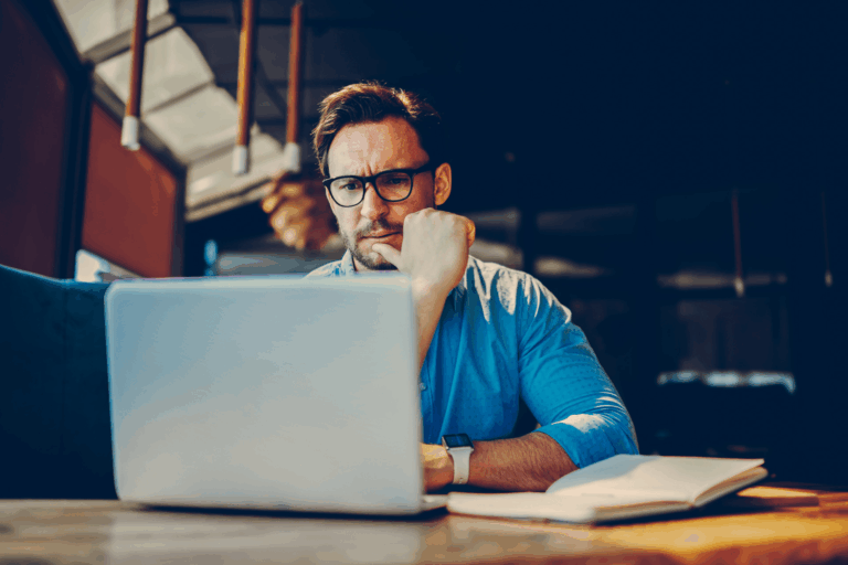 Man in blue shirt and glasses thinking while looking at laptop, considering questions for marketing agency.