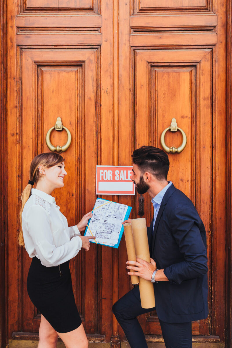 Real estate agents discuss property plans outside a "For Sale" door.